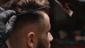 Professional male barber in black rubber gloves making haircut of handsome bearded man with sharp razor at berbershop. Concept of stylish hairdressing and shaving.