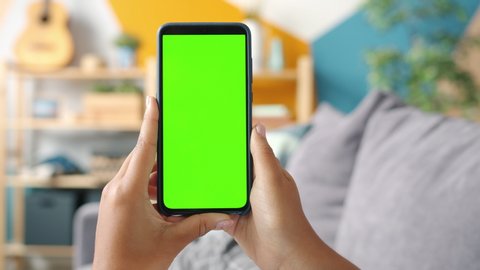 Close-up shot of green screen template smartphone in female hands at home, girl is watcing content without touching gadget screen. Modent technology and information concept.
