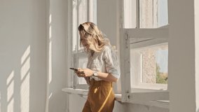A pretty smiling woman with blond hair is using her smartphone with wired headphones in white office near open windows