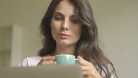 A pretty positive young woman is using laptop computer while drinking coffee indoors