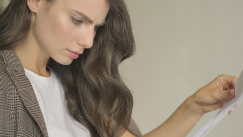 A serious concentrated young woman is working with documents while sitting at the table with a laptop indoors