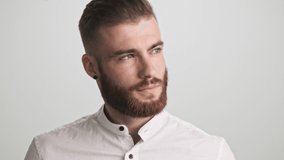 A serious bearded man wearing white shirt is touching his chin while thinking about something isolated over white wall background