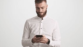 A positive calm young bearded man wearing white shirt is using a smartphone isolated over white wall background