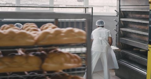 Process of load the raw bread in the oven machine of two bakery workers , walking around in front of the camera the baker in a special uniform and other professional worker transported the shelves of