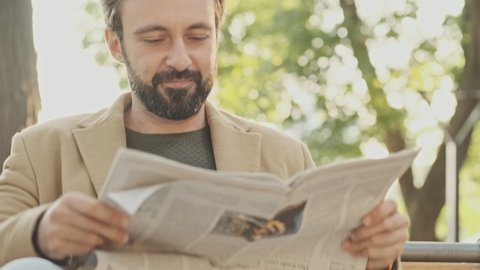 Cheerful elegant bearded man in coat reading newspaper while sitting in the park outdoors