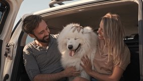 An attractive couple man and woman are petting a dog while sitting in the car trunk