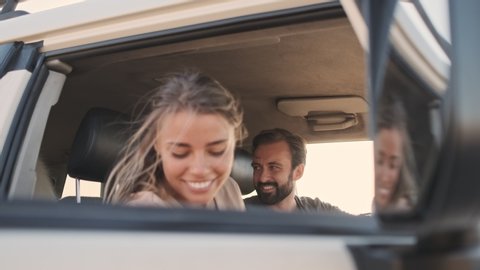 A smiling young woman sits in the car with her boyfriend and a dog in the back seat outdoors in summer