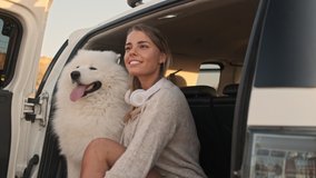A smiling positive young woman is sitting with a dog in the trunk outdoors in summer