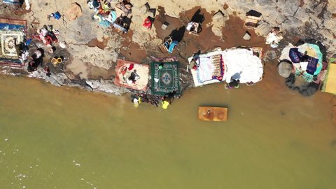 The drone flies up towards the left with a topdown down view upon a busy rural riverside with people washing their clothes in the river with a local village in the background in Mali Aerial Drone 4K