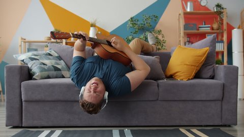 Joyful guy is playing the guitar singing wearing headphones having fun on sofa at home. Modern entertainment, music and happy young people concept.