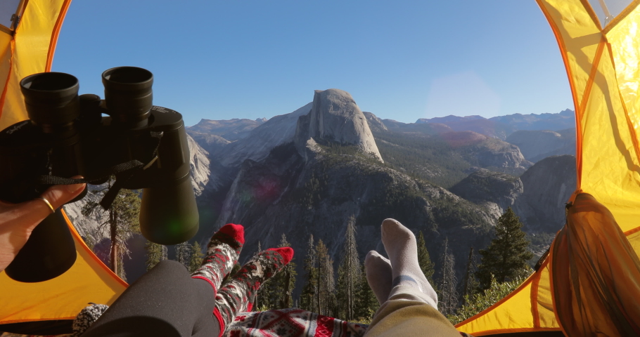 A couple, having a rest in Yosemite National Park in California, USA, lays inside the camping tent with the view to the Half Dome rock. One person gives the big black binoculars to the other. 4K