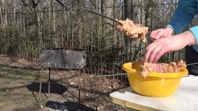 man hands take raw spiced meat from yellow dish and put on metal spit. Fire flame burn in rusty barbecue in back yard. Male in blue jumper. 4K UHD video clip.
