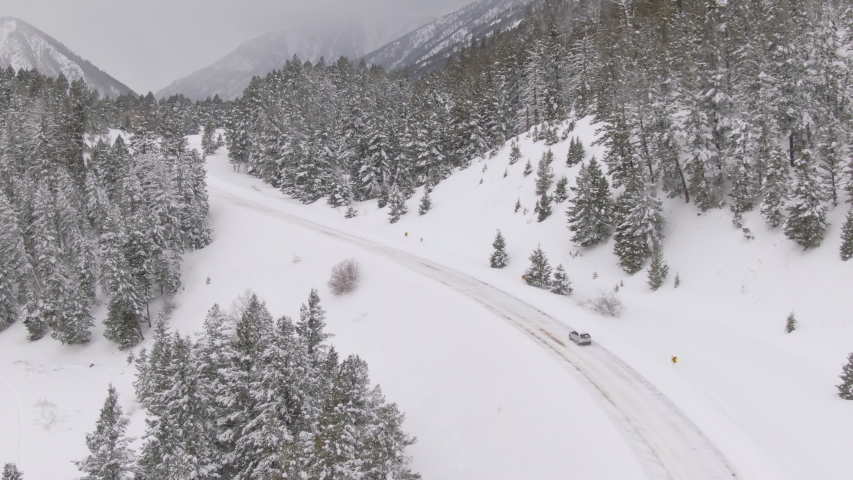 DRONE: Commuter in car making their way down a snowy road in Spokane, Washington. Flying behind a car driving down a snow covered road leading through the dense pine forest in the scenic US mountains.