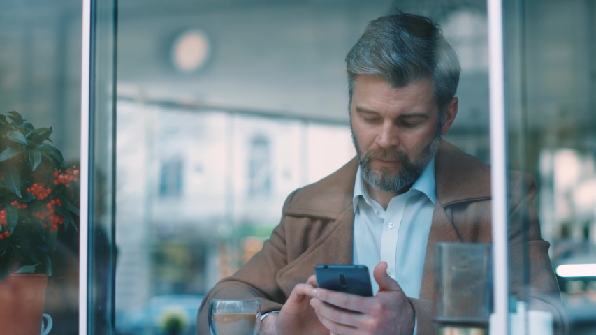 View through the window of attractive serious middle aged businessman using phone at coffee shop texting message  | Shutterstock HD Video #1044817183