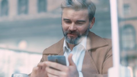 Close up rotation view of gorgeous middle aged bearded business man with beige coat uses phone drinks coffee at cafe texting message working successful people relaxing new technologies communicating