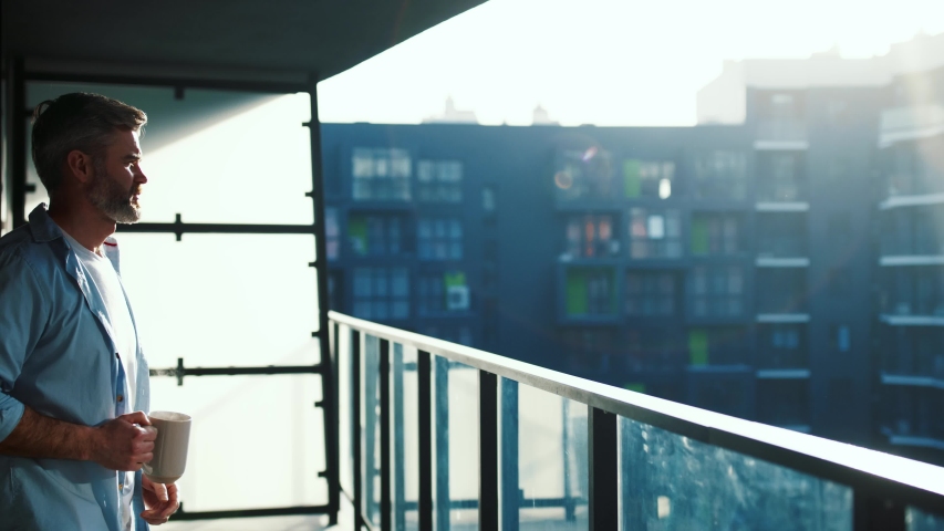 Handsome caucasian middle aged man stands in the balcony with great views and drinks hot coffee or tea, sun shine, enjoying beautiful morning feeling happy Royalty-Free Stock Footage #1044817213