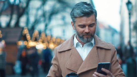 Close up view of handsome mature caucasian man walks down the city center street, drinks coffee and uses phone, texting message, feels happy, city lights on background slow motion