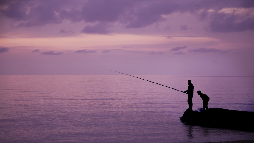 Gimbal shot of man and a child fishing on background of sea at sunset in evening time. Two people fishing on the cliff near the river at violet sunset