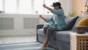Afro-American lady in vr glasses is enjoying driving game at home moving arms and legs indoors relaxing alone with cool innovation. Entertainment and modern devices concept.