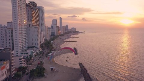 Aerial Drone Shot of Motorized Paraglider/Paramotor flying over Sand Beach of Bocagrande, Cartagena, Colombia next to lots of Skyscrapers & High Rise Buildings into orange Sunset at Caribbean Coast.