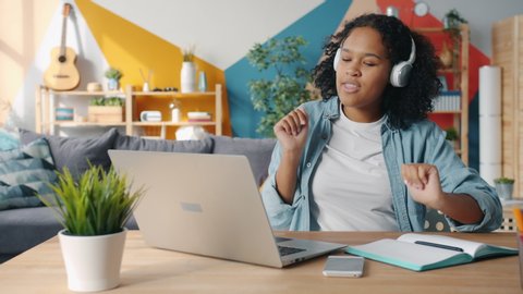 Joyful Afro-American female student in headphones is using laptop at home dancing having fun alone. Modern technology, youth and emotions concept.