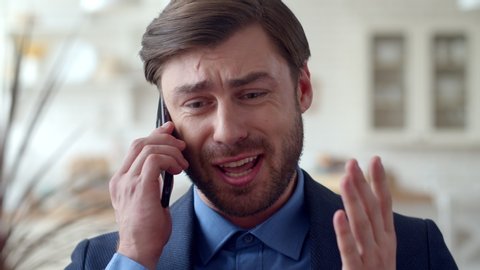 Closeup angry businessman talking mobile phone emotionally at home interior. Stressed guy making phone call in slow motion. Portrait of serious man gesturing with cell phone at remote workplace.