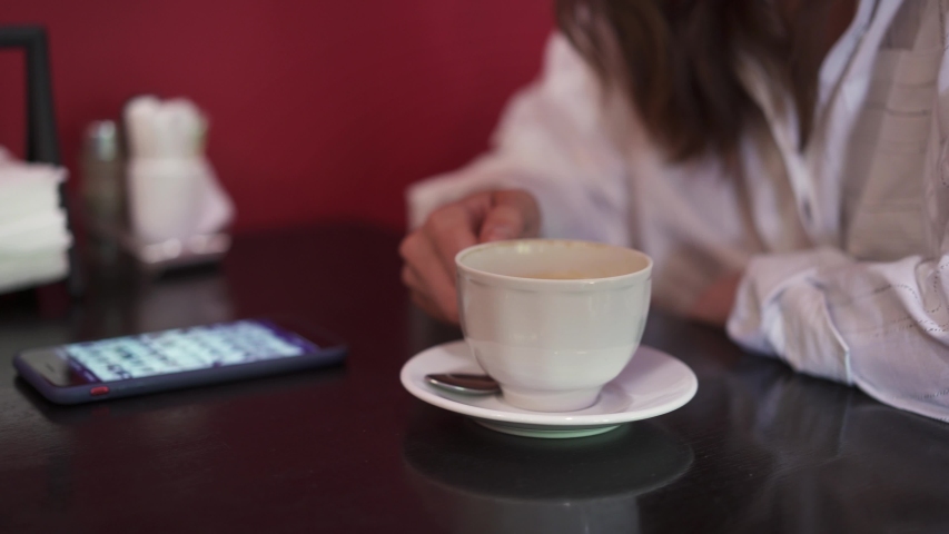 Close up profile shot of young female drinking a cappuccino from a white cup in a cafe and scrolling the phone. Young woman, no face, drinking coffee