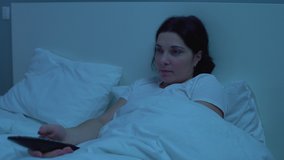 Young female switching tv channels with remote controller, lying in bed alone