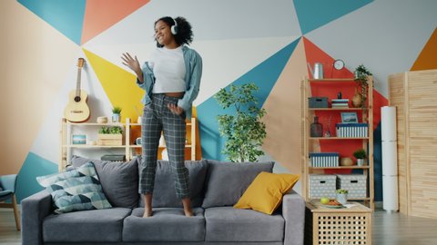 Mixed race woman in headphones is dancing on sofa in apartment having fun alone feeling happy and carefree. Joyful young people and entertainment concept.