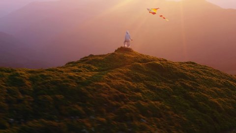 Happy woman flying a rainbow kite on mountain hill, observing magnificent nature scenery and summer sunset. Cinematic landscape. Concept of freedom.