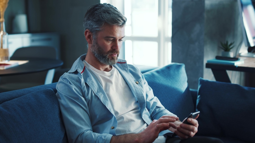 Close up view of cheerful middle aged man with grey hair sitting on the sofa using phone smile in the modern apartment texting message scrolling tapping technology isolated lifestyle slow motion Royalty-Free Stock Footage #1044844771