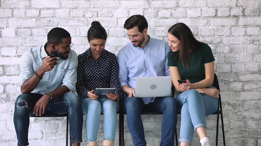 Happy diverse young adult business people group sharing social media news using gadgets, multiethnic friends holding devices and talking sit on chairs in row, technology obsession addiction concept Royalty-Free Stock Footage #1044846376