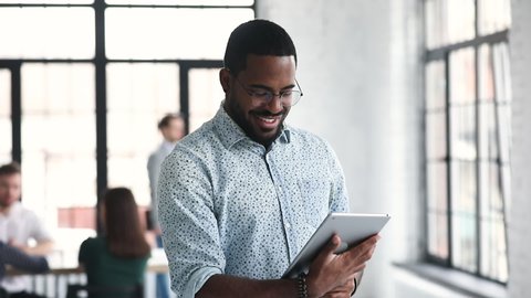 Young professional african businessman worker using digital tablet corporate software standing in office, smiling ethnic male manager working online on pad computer tech in modern company office