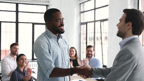 Proud confident african american male professional get promoted handshake boss pleased by multiracial team feedback applause, employee respect, recognition at work, staff reward concept, slow motion