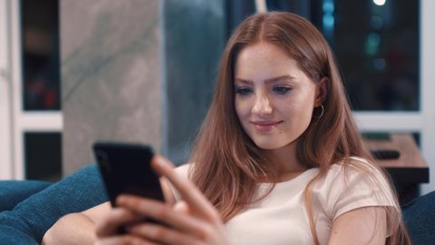 Close up view of pretty redhair young woman sitting lying on blue coach uses phone and smiles at modern room Texting sharing messages on social media enjoying feel happy mobile technology evening