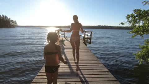 A young slender woman with daughter running on a wooden pier and jumping into the lake after the sauna in summer white night in Finland.