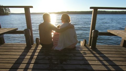 Couple with child sitting on a wooden jetty at sunset near the lake at sunset after the sauna in summer white night in Finland.