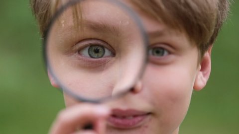 Closeup view of cute white young kid holding magnifier glass in hand and looking at camera through it smiling cheerfully. Real time full hd video footage.