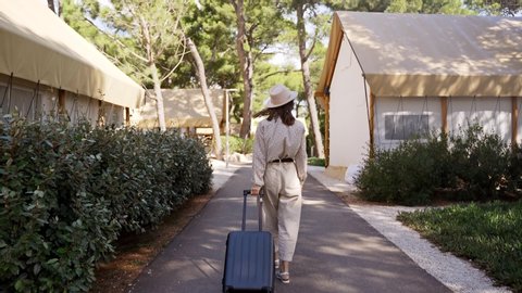 young beautiful girl in beige pants and a light shirt is walking with a suitcase along the path in glamping. The view from the back. Video de stock