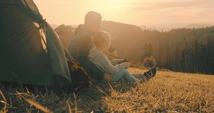 Happy traveler father and son sitting on grass and look through binoculars, bonding around mountain under sun light enjoying the leisure and freedom. 4K slow motion video