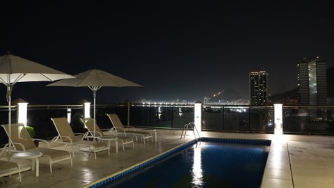Rio de Janeiro, Brazil - January 17, 2020: Night timelapse of luxury and trendy hotel rooftop with swimming pool, umbrella and long chairs. City light and skyscraper buildings visible in the backgroun