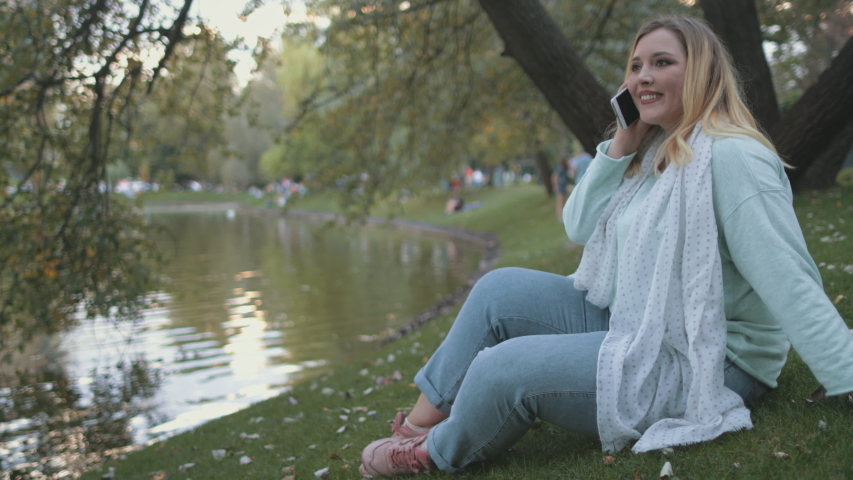 Woman Plus Size Sit On Lake Bank Park Background. Pretty Caucasian Girl Talk Smartphone Summertime City Parkland River Surface. Happy Lady Smile Enjoy Mobile Conversation Slow Motion 4K
