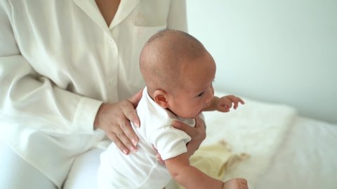 From above of mom patting adorable newborn son massaging back of Asian baby on bed at home