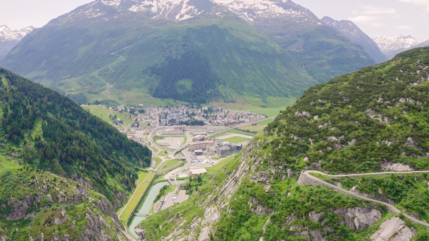 Switzerland. The Gotthard Pass near Andermatt, a historic mountain pass in the Swiss Alps, is shown in a sweeping aerial view that reveals a winding road and tunnel carved through a dramatic alpine landscape. The scene captures the rugged mountain slopes covered in dense green forest and rocky outcrops, with a small town nestled in the valley below and snow-capped peaks rising in the background under a clear summer sky. The pass has served as a vital north-south corridor since the 13th century, when the first route across the Schollenen Gorge opened, and remains a powerful symbol of Swiss engineering and alpine transit. The camera glides above the terrain, emphasizing the scale of the mountains and the integration of the road and tunnel infrastructure into the natural environment. The atmosphere is serene and majestic, with bright daylight illuminating the lush green valleys and the stark rocky cliffs.
