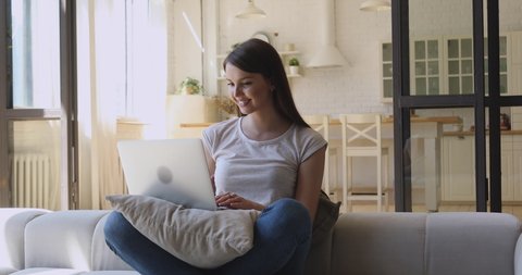 Happy young woman relaxing on sofa with computer, chatting with friends online in social network, answering messages emails, web surfing information, shopping in internet store in cozy studio home.