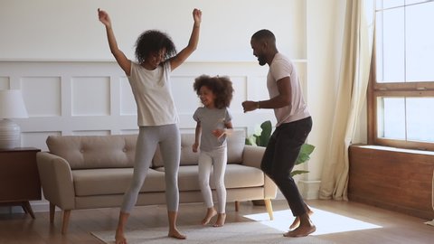 Overjoyed smiling african ethnicity family couple dancing with cute daughter at living room. Happy mixed race family having fun, involved in funny activity together on weekend morning at home.