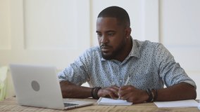 Concentrated young african ethnicity guy sitting at table, watching educational webinar, writing down notes, rewinding video. Focused mixed race male freelancer working remotely at home or office.