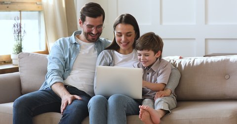 Happy family young parents couple and cute preschool kid son using laptop looking at computer screen enjoying watching funny social media video doing online shopping relaxing on sofa at home together