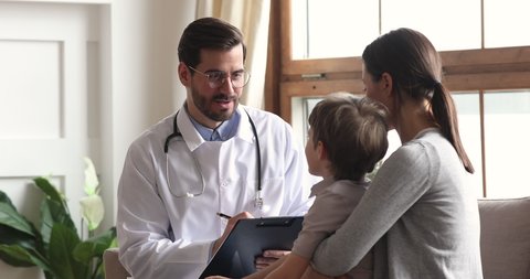 Smiling friendly male pediatrician wear white coat hold clipboard talk to preschool kid boy patient do pediatric checkup give high five, child with mother visit doctor sit on sofa in medical office