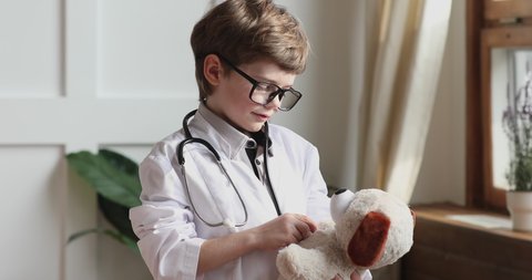 Funny small preschooler boy wear medical coat glasses hold stethoscope pretend listen toy dog patient, cute adorable little smart child play hospital game as vet doctor, children veterinary concept